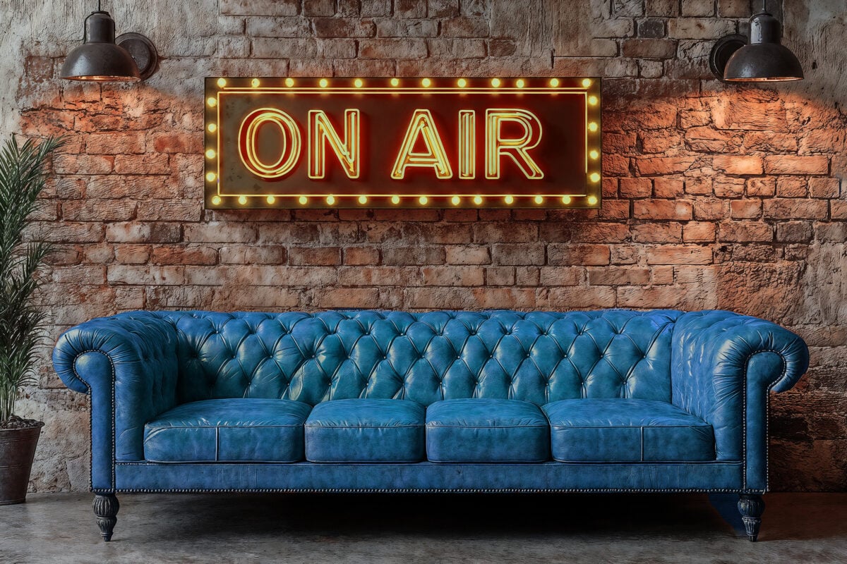 A vintage red leather sofa lit by spotlights with a brick wall backdrop. A neon sign reading “On Air”. For live broadcasts.