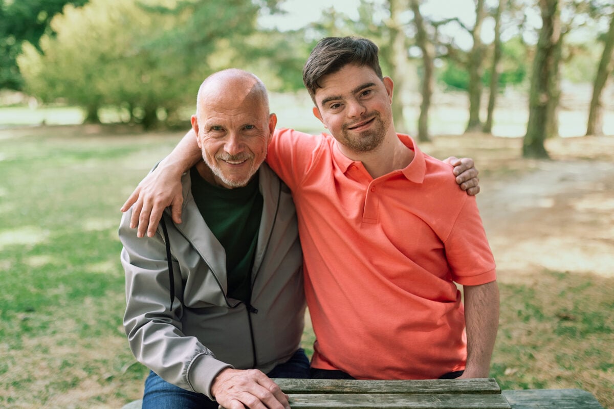 A happy senior father with his young son with Down syndrome embracing and sitting in park.
