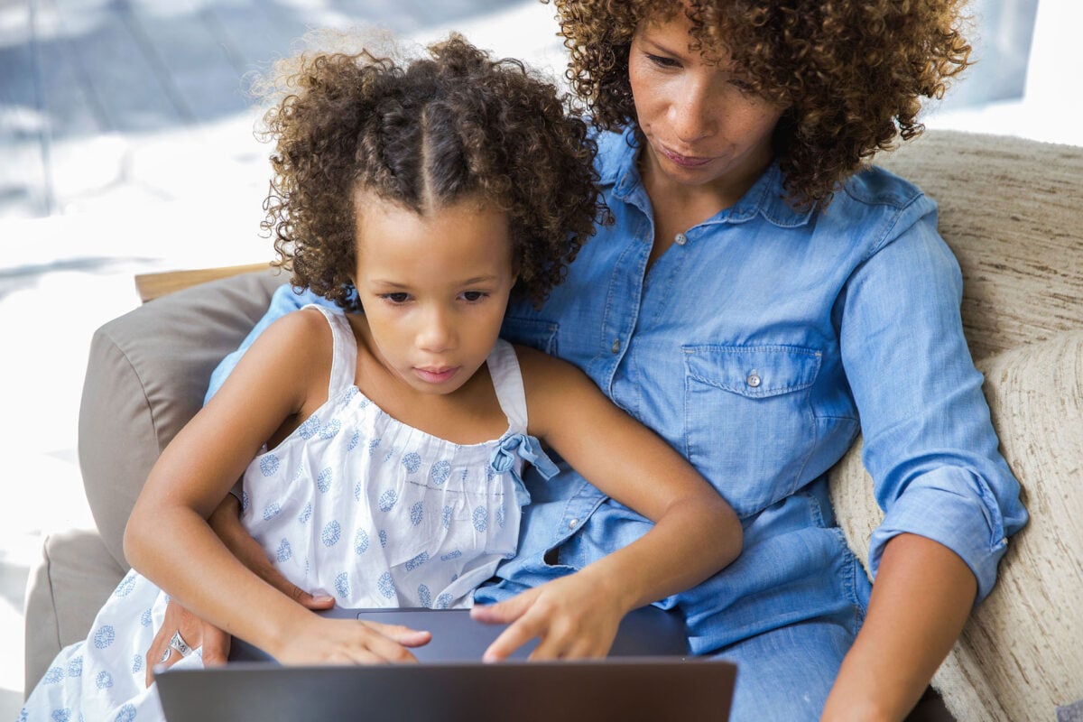 A mom is sitting on the couch with her young daughter. They are using the internet together at home.