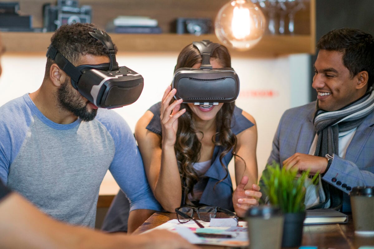 A multi-ethnic group of adult business-people are indoors in a board room. Some are wearing formal clothing while others are wearing casual clothing. Two people are trying virtual reality headsets while their coworkers watch.