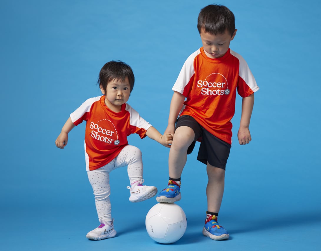 Two kids in soccershots uniform stepping on a soccer ball.